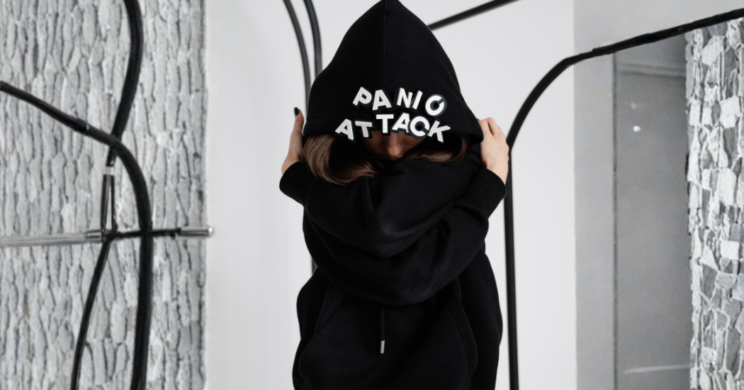 Γυναίκα με κουκούλα που γράφει panic attack, αγκαλιάζει τον εαυτό της. κρίση πανικού συμπτώματα